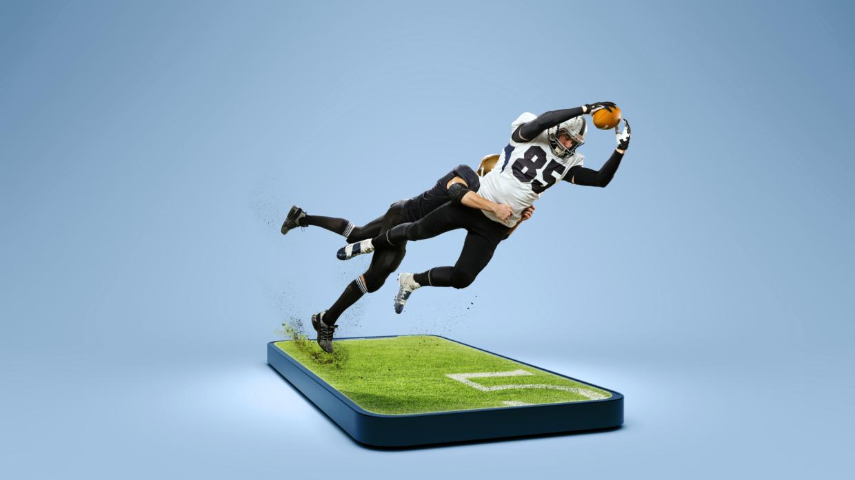 two football players dive for a ball on top of a supersized phone, whose screen appears to be grass