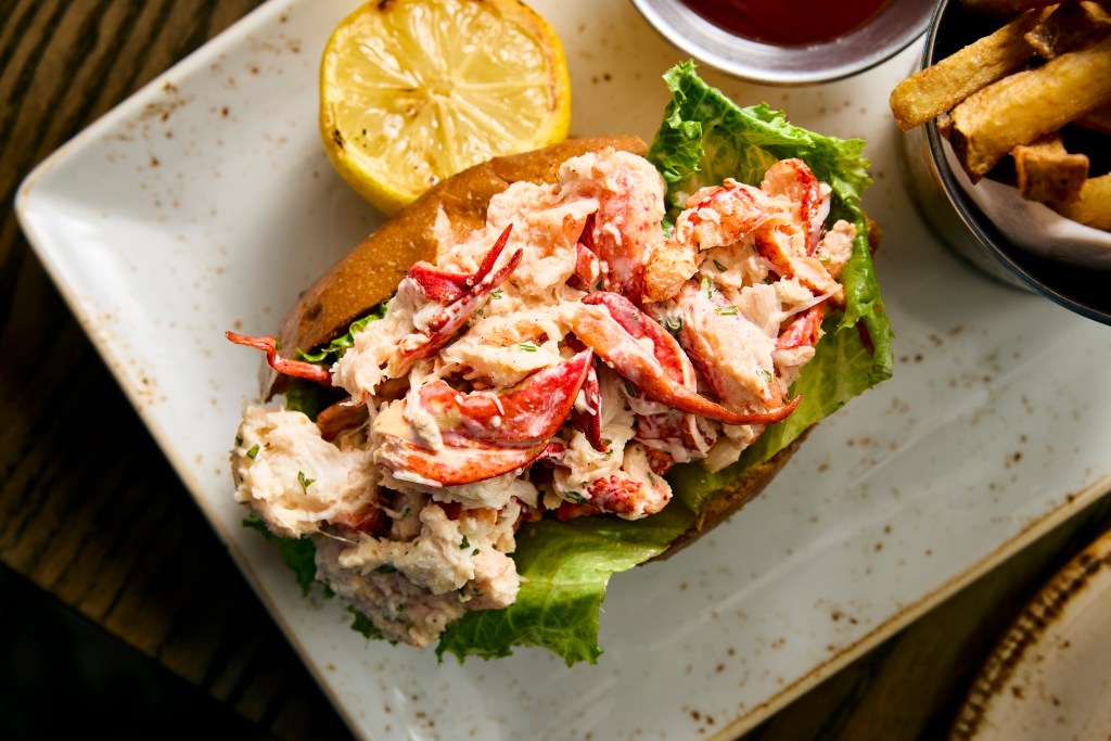 A lobster roll on a plate with fries and a grilled lemon half.