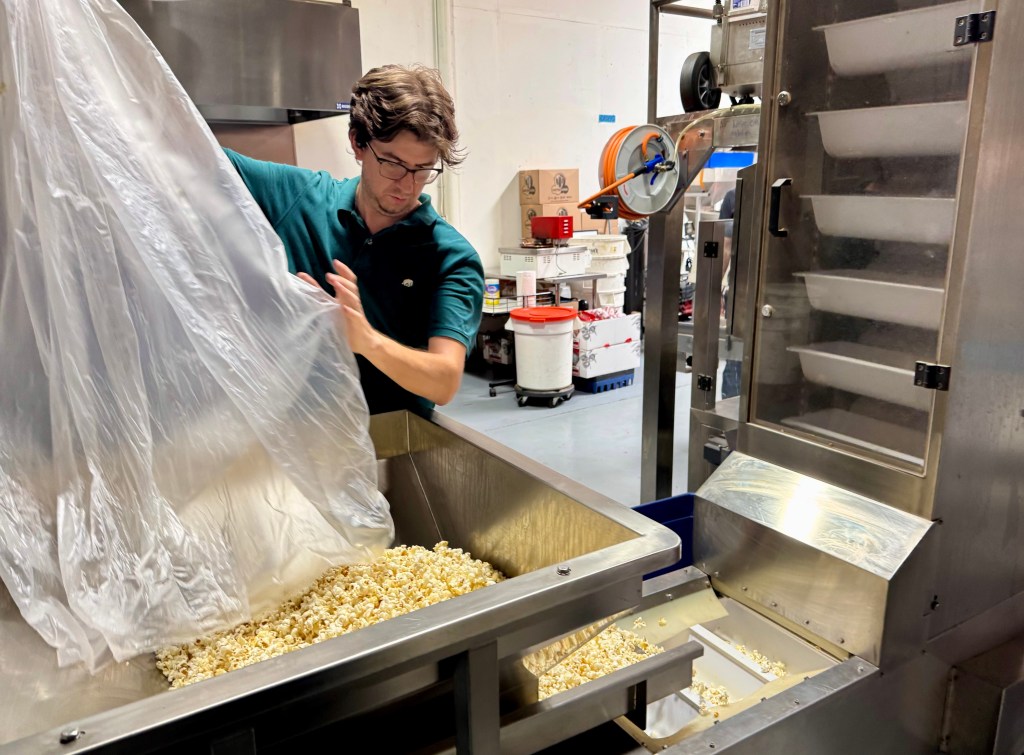 A man pours a large bag of popcorn into the hopper of a machine that will package it into individual bags.