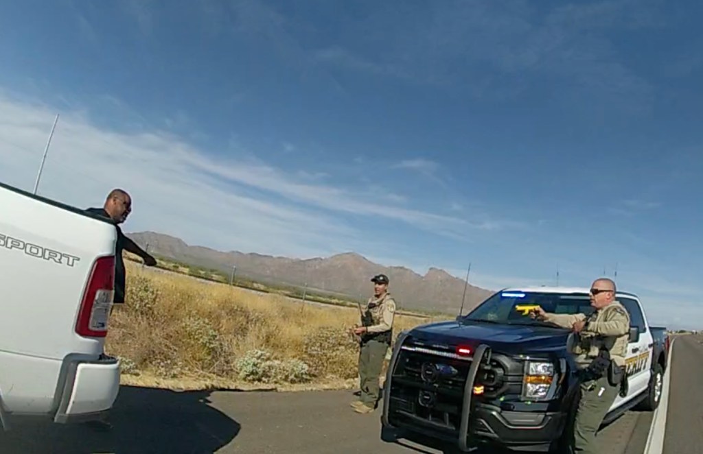 at a roadside traffic stop a sheriff's deputy aims a taser at a man standing with arms out several feet away