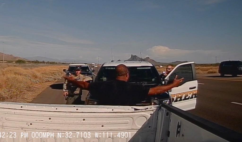 a man at the back of a pickup truck bed stands with his arms out, as two sheriff's deputies yell at him on the side of a highway