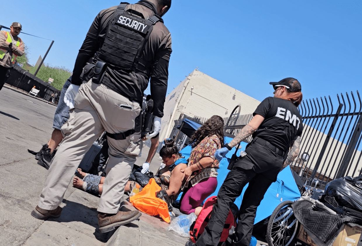 a security guard and an e.m.t. stand above a woman slumped on a sidewalk curb on a bright day
