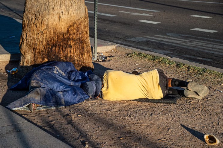 a unhoused man in a yellow shirt laying, back to the camera, at the base of a tree