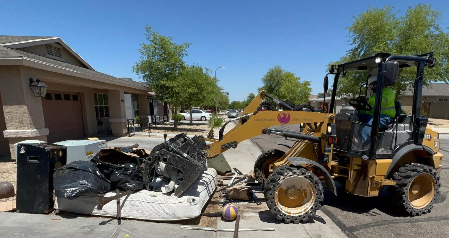 A City of Phoenix backhoe loader scoops up debris on a residential curb.