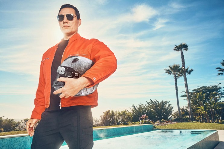 A man in an orange jacket holding a motorcycle helmet.