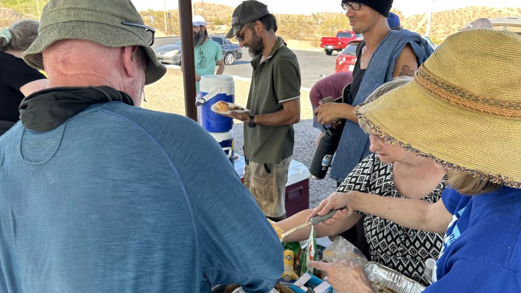 people crowd around food on a picnic table