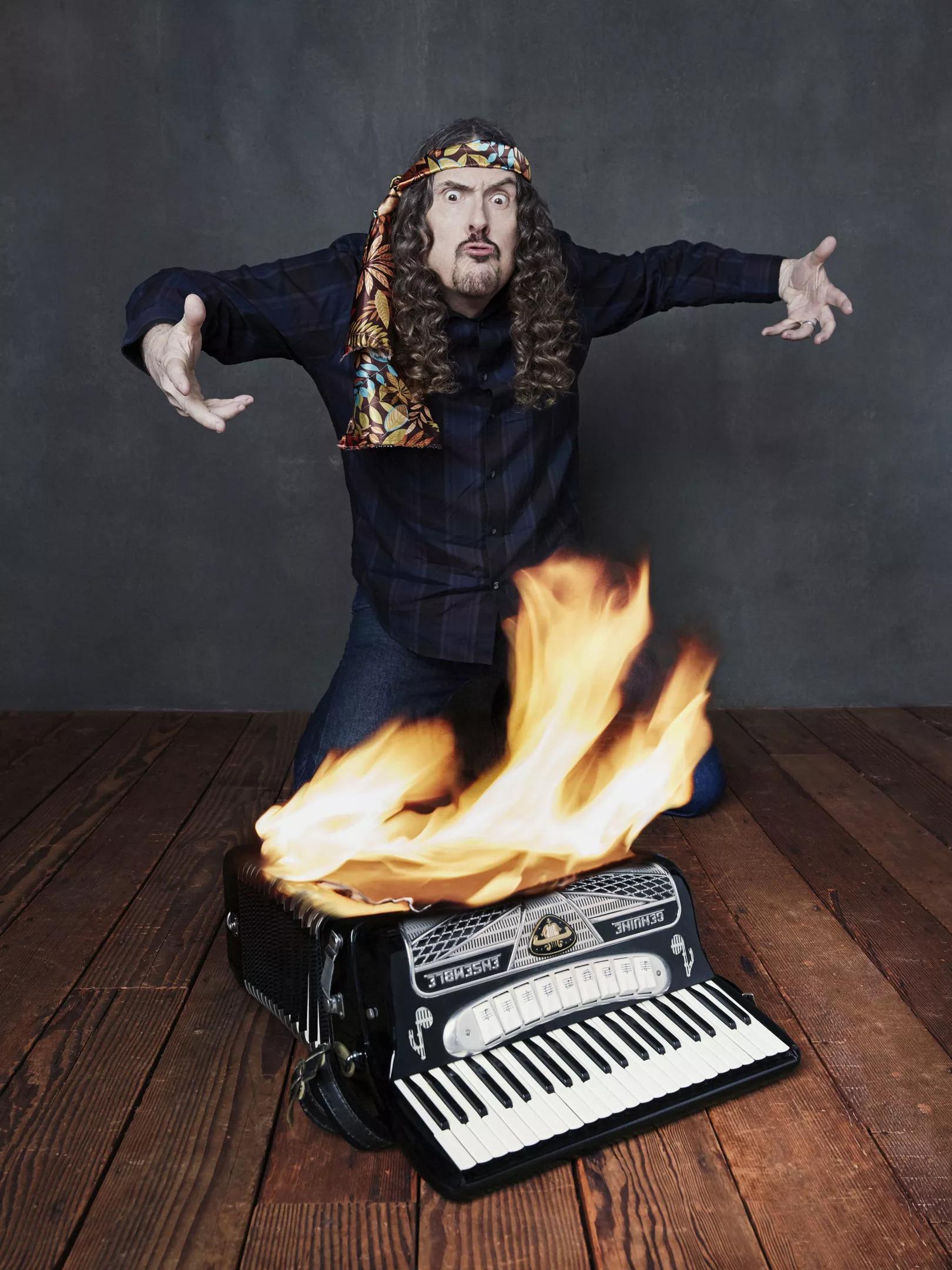 A long-haired man standing over a burning accordion.