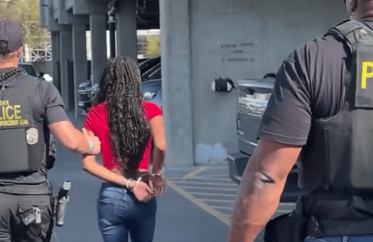 seen from behind, two police officers escort a handcuffed young black girl with braids