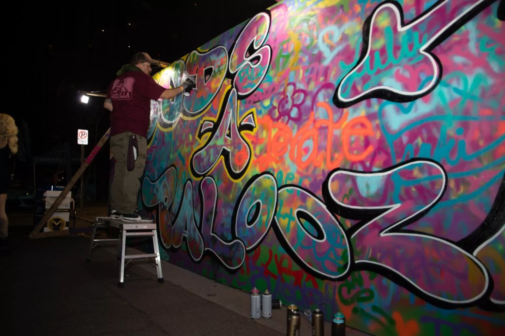 A man spraypaints on a wall that says "buds-a-palooza"