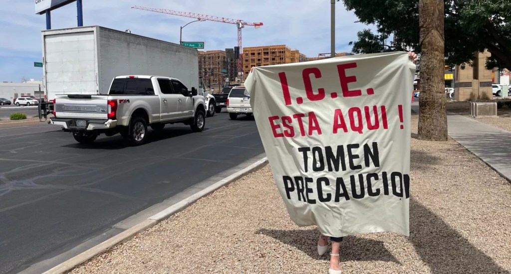 A woman holds up a large banner that reads: “ICE is here! Take precautions,” in Spanish at the Phoenix Immigration Court in Phoenix
